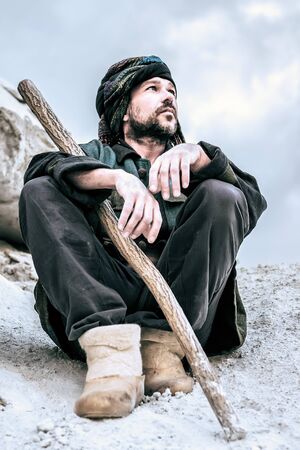 Man in the patterned turban with a stick sitting and seriously looking to the side on a light...