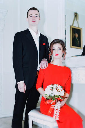Portrait of a loving couple in a light studio - a woman in a red dress with a bouquet sits, a man in a suit stands. Soft Focus Filter.の写真素材