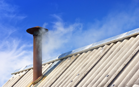 Rusty chimney pipe on a slate covered roof of an old house. White Smoke from the chimney against the blue sky with clouds. Abstract concept background with space for copy, selective focus.の写真素材