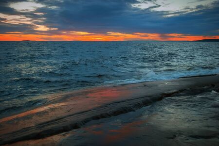Orange sunset in dramatic sky over a stormy lake with a rocky shore. Lake Onega, Karelia, Russia. Selective focus, blur filter.の写真素材
