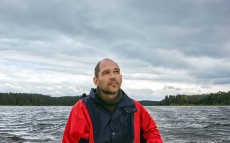 A pensive bearded mature man in a storm jacket looks at a stormy cloudy sky over a wavy forest lake. Selective focus in the foreground, blurred natural background with space for copy.の写真素材