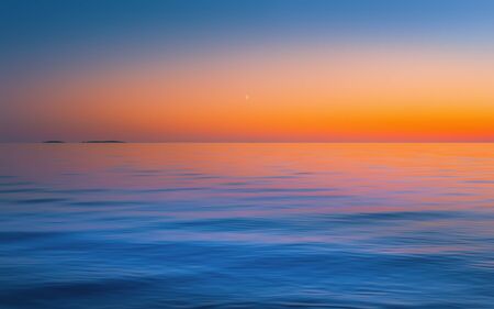 The crescent moon in a clear sky after red yellow sunset reflected in the flowing water surface. Abstract motion blurred seascape background  in orange blue colors with copy space. Russia, Lake Onega.の写真素材