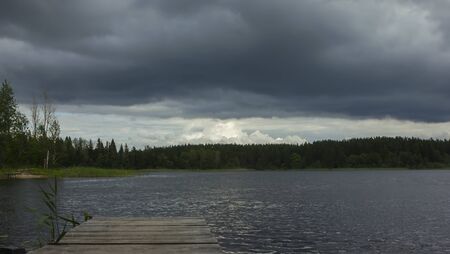Dramatic dark stormy sky with huge heavy rain clouds over the forest lake in windy weather. Atmospheric landscape with space for copy, selective focus. Seliger Lake, Russia.の写真素材