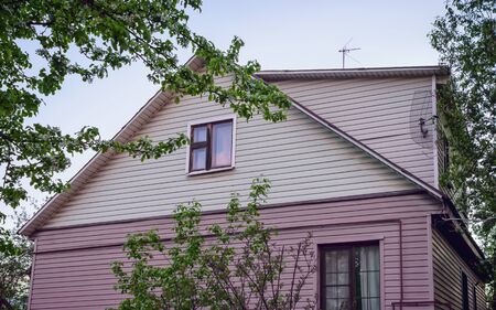 Low angle view of the top of a country house with an attic and a mezzanine. Simple residential cottage with siding paneling in the spring garden. Russia. Selective focus.の写真素材