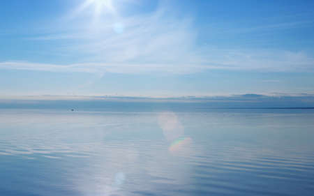 Blue sky reflected in a smooth lake surface. Water glares and shines in the rays of the evening sun with lens flare. Summer seascape background with space for copy. Russia, Vologda Oblast, Lake Beloye.の写真素材