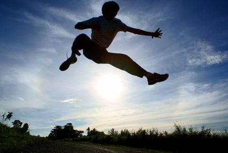 Silhouette of person jumping over sun with blue sky and cloudsの写真素材