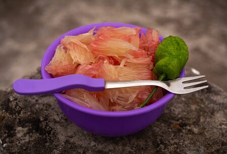 POMELO (ROBAB TENGA) CITRUS FRUIT INNER PARTS WITH GHOST CHILI PEPPER (BHUT JOLOKIA) AND FORK SPOON IN A VIOLET PLASTIC BOWL PLACED ON GREY SURFACE PILLARの写真素材