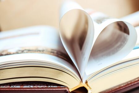 Gold pages of a book curved into a heart shape on wooden blurred background.