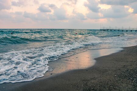 Sea surf waves roll onto the beach and bring seaweed. An off shore wind blows the white foam top back over the wave. Bright turquoise water and cloudy sky. Mideterranean sea.の写真素材