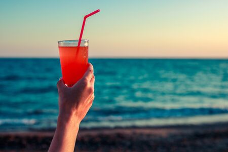 Female hand holds a glass with a delicious cocktail on a background of sea and beach. Beach party...