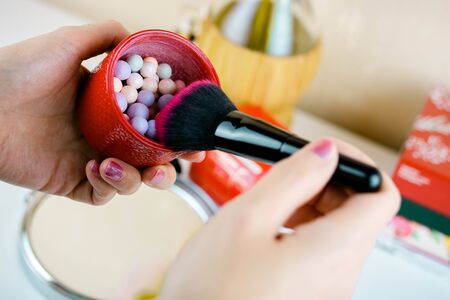 Female hand holds multicoloured highlighting round powder peals in round red box. Makeup product for face treatment. Ready to apply powder with a brush.の写真素材