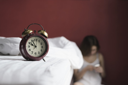 Retro alarm clock on the bed with woman sitting strain and using phone,unhappy In the corner of the room, の写真素材