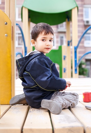 Serious cute boy sits on playground with houses on backgroundの写真素材