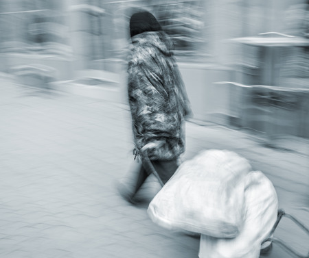 An elderly woman carries bags down the street on a trolley.  Intentional motion blurの写真素材
