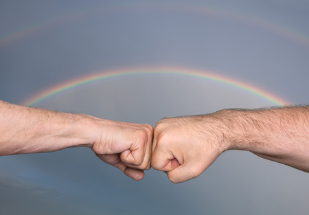 Two men bumping fists against the background of a stormy sky with rainbowの写真素材