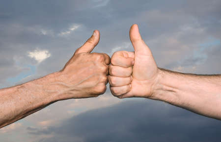 Friendship concept. Two men bumping fists with thumbs up against sky with cloudsの写真素材