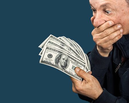 Confused elderly man with fan of dollars on dark blue background with copy-spaceの写真素材