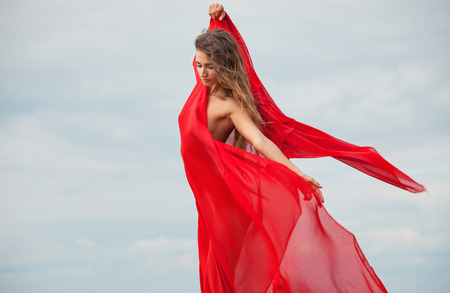 Beautiful nude woman with red fabric posing on sea beach against sky backgroundの写真素材