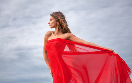 Beautiful young woman with red fabric posing against sky backgroundの写真素材