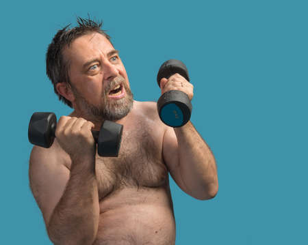 An elderly fat and wet man exercising with dumbbells isolated on blue with copyspaceの写真素材