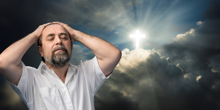 Religious conversion concept. Elderly man thinking about faith and God. Portrait against the sky with a glowing cross symbol of faith.の写真素材
