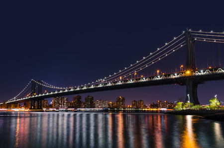 Manhattan Skyline and Manhattan Bridge at night with reflections in East River. Manhattan Bridge is...