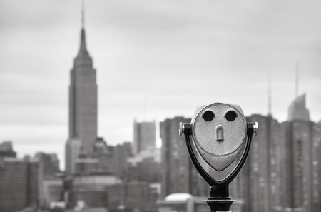 Black and white image of an old binoculars and New York City Manhattan skyline with the Empire State Building over East River viewed from Brooklyn on an overcast dayの写真素材