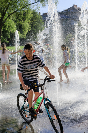 UZHGOROD, UKRAINE - May 28, 2017: Happy children playing in a water fountain in a hot day. A boy is riding a bicycle in a fountainのeditorial素材