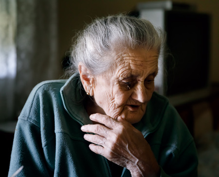 Old depressed woman. An elderly lonely woman sits at a table in the kitchen near the windowの写真素材