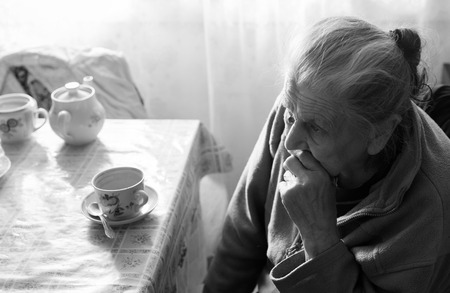 Old depressed woman. Black and white image of an elderly lonely woman sits at a table in the kitchen near the window and drinking tea.の写真素材