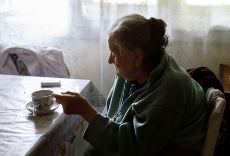 Old depressed woman. An elderly lonely woman sits at a table in the kitchen near the window and...