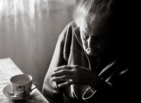 Black and white portrait of a very old tired woman sitting near the window in the kitchen of her house の写真素材