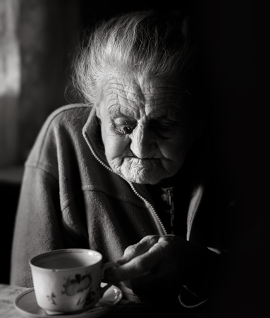 Black and white portrait of a very old tired woman sitting near the window in the kitchen of her...
