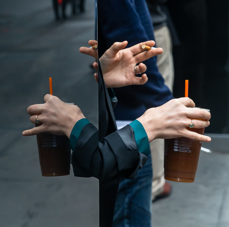 New York, USA - May 03, 2016: Coffee break in the streets of New York City. Hand of a woman with a cup of coffee and the hand of a man with a cigarette reflected in a glass caseのeditorial素材