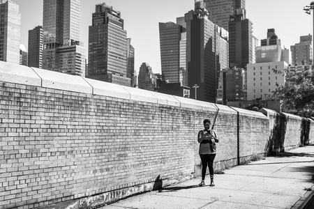New York, USA - Sep 23, 2017: Manhattan street scene. Black young woman with a mobile phone is standing near yellow brick fence on the streets of Manhattan in New York Cityのeditorial素材