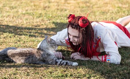 Young woman with scarlet dreadlocks in national dress lying on the grass and playing with the cat. Outdoors portraitの写真素材