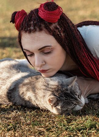Young woman with scarlet dreadlocks in national dress lying on the grass and playing with the cat. Outdoors portraitの写真素材