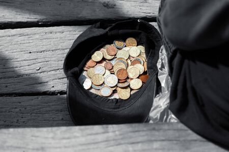 A beggar sits near a cap with coins and asks for alms. Cap with a few alms coins