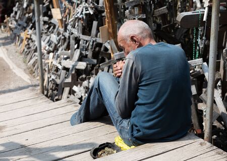 SIAULIAI, LITHUANIA - JUL 22, 2018: A beggar on a mountain of crosses asks for alms. A beggar sits near a cap with coins and asks for alms. Cap with a few alms coinsのeditorial素材