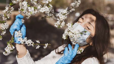 Lifestyle in the new environment concept. Spring and the coronavirus epidemic. A young woman in medical gloves and a protective mask enjoys spring flowering treesの写真素材