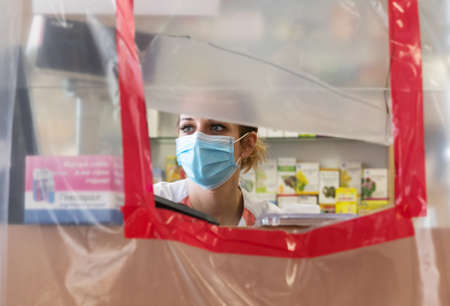 Bucha, Ukraine - Apr. 24, 2020: Coronavirus pandemic. Demarcation the areas with the film during quarantine in Ukrainian pharmacies. Young woman pharmacist in a medical maskのeditorial素材