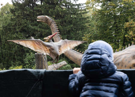 Two-year-old boy in a dinosaur park looks from behind the fence at a group of dinosaursの写真素材