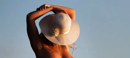 Young nude woman with a white hat standing on a stone by the sea in the rays of the sunset sunの写真素材