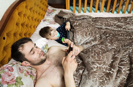 Happy family concept. A young father and his little son are lying in bed and playing with mobile...