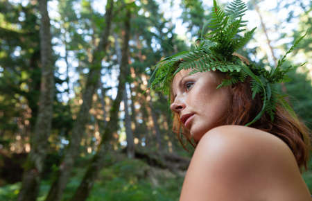 A beautiful young nude woman with fern wreath on her head enjoying nature in the forest riverの写真素材