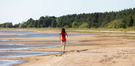 Beautiful girl outdoors enjoying nature. Young woman with scarlet dreadlocks in a red bathing suit walks along the sand towards the seaの写真素材