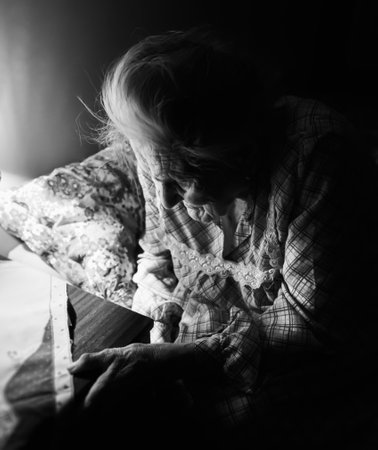 Old woman sitting on sofa in dark room near coffee table with a light from table lampの写真素材