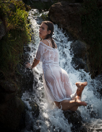 Seminude young woman in a white dress enjoys the freshness and coolness in the water jets of the waterfallの写真素材