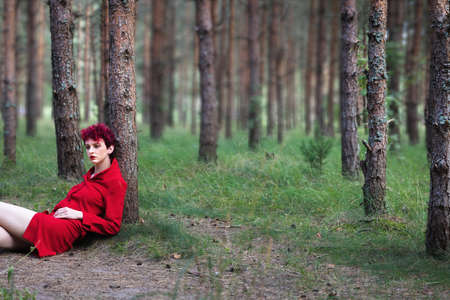 Youth and beauty concept. Young woman with pink hair and in a red dress posing in a pine forestの写真素材