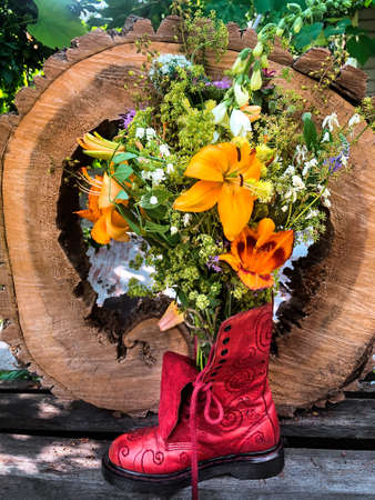 Romantic bouquet of farm flowers in a red boot vase against background of a cut of an old tree. The composition includes flowers lily, daylily, foxglove, parsley, parsnips, clematis, geyhera, monardaの写真素材